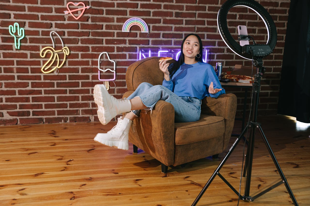 about-02 Young Asian woman influencer streaming live video while enjoying pizza in a stylish room with neon décor.